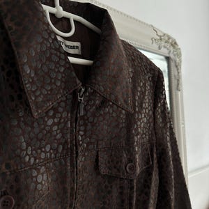 May include: A brown jacket with a textured pattern, featuring a zipper closure and two front pockets. The jacket is hanging on a white hanger. The label reads "RY WEBER".