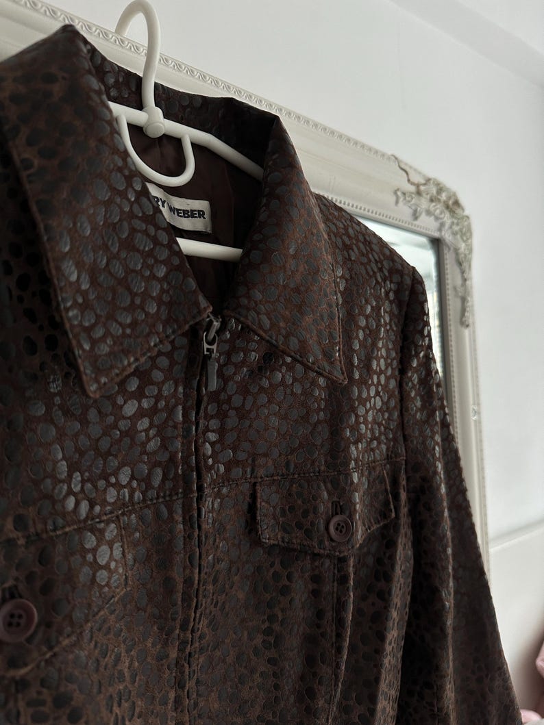May include: A brown jacket with a textured pattern, featuring a zipper closure and two front pockets. The jacket is hanging on a white hanger. The label reads "RY WEBER".
