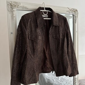May include: A brown leather jacket with a textured, spotted pattern hangs on a white hanger. The jacket has a collared neckline, two front pockets, and a zipper closure. The brand name "Gerry Weber" is visible on the hanger.