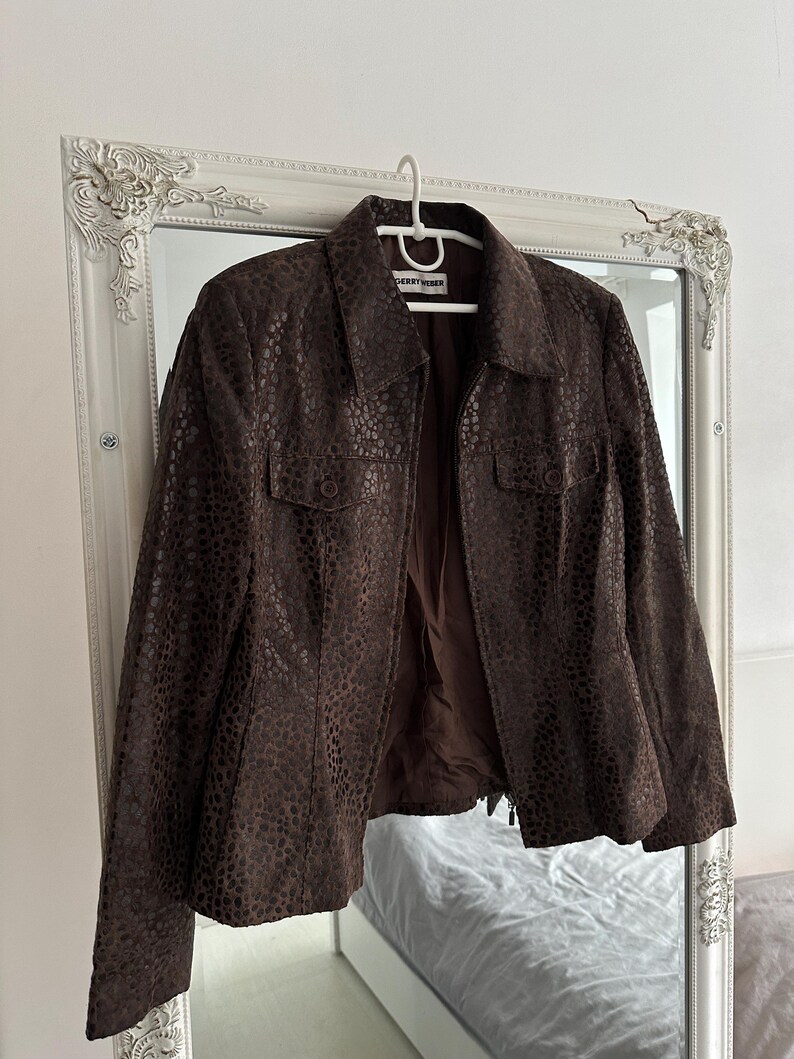 May include: A brown leather jacket with a textured, spotted pattern hangs on a white hanger. The jacket has a collared neckline, two front pockets, and a zipper closure. The brand name "Gerry Weber" is visible on the hanger.