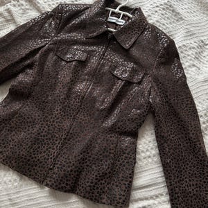 May include: A brown jacket with a spotted pattern, featuring a collar, two chest pockets, and a zipper closure. The jacket is hanging on a white hanger.
