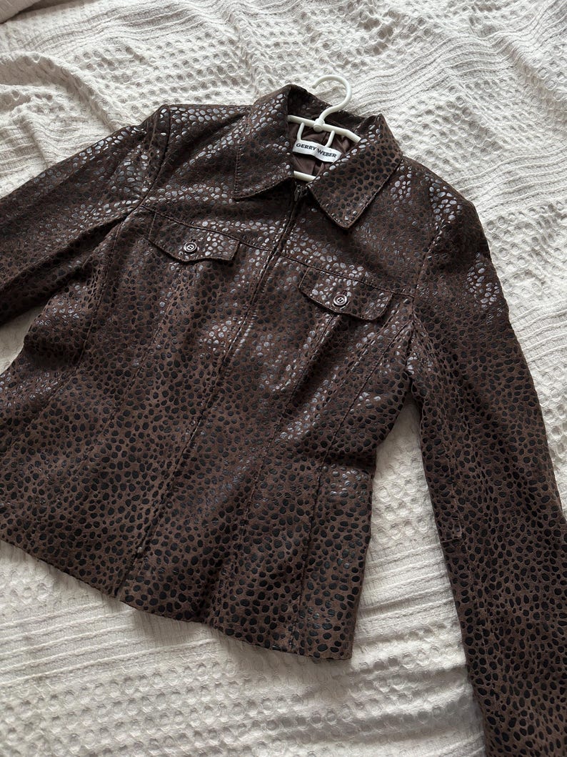 May include: A brown jacket with a spotted pattern, featuring a collar, two chest pockets, and a zipper closure. The jacket is hanging on a white hanger.