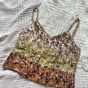 Pode incluir: Um top de camisola com lantejoulas com decote em V e alças finas em forma de corrente. A parte superior faz a transição de lantejoulas douradas para verdes e castanhas. A parte superior está sobre uma superfície branca texturizada com uma peça de crochê branca no fundo.