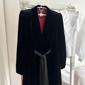 以下が含まれることがあります： ノッチドラペル、長袖、ウエストに同色の黒いサテンの結びベルトが付いた黒いベルベットのブレザー。内側の裏地は、控えめなドット柄の濃い赤色の生地です。首元には白いブランドラベルが見えます。