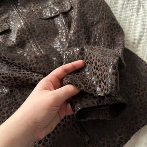 May include: Brown jacket with a spotted pattern. The jacket has a collared neckline, two chest pockets, and long sleeves. The fabric appears to be leather or a leather-like material.