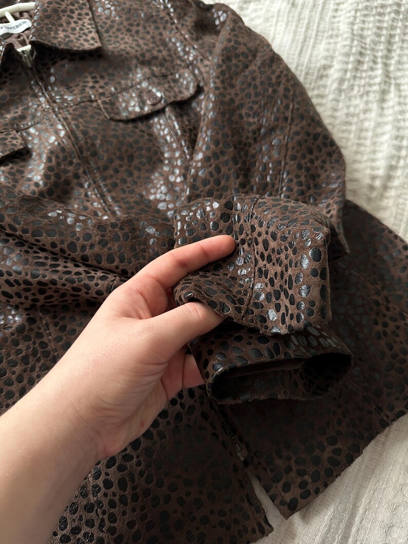 May include: Brown jacket with a spotted pattern. The jacket has a collared neckline, two chest pockets, and long sleeves. The fabric appears to be leather or a leather-like material.