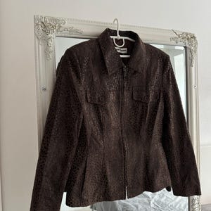 May include: A brown, textured jacket with a collared neckline and a zipper closure. The jacket features two small pockets on the chest and is hanging on a white hanger. The jacket is displayed in front of a decorative mirror.