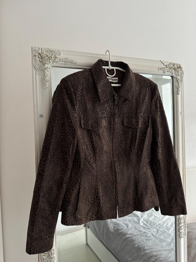 May include: A brown, textured jacket with a collared neckline and a zipper closure. The jacket features two small pockets on the chest and is hanging on a white hanger. The jacket is displayed in front of a decorative mirror.