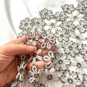 May include: A white beaded floral lace with dark gray accents. The delicate, openwork design features repeating flower patterns, creating a decorative textile. The lace is held in a hand, showcasing the intricate beadwork.