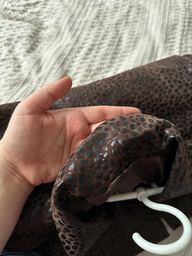 May include: Brown jacket with a dark spotted pattern. The jacket is hanging on a white hanger. The interior lining is a solid brown color.