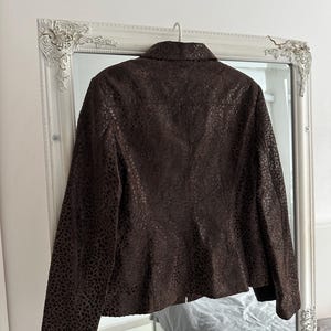 May include: A brown leather jacket with a textured, spotted pattern hangs on a hanger in front of a mirror. The jacket has a collared neckline and long sleeves. The mirror has a white ornate frame.