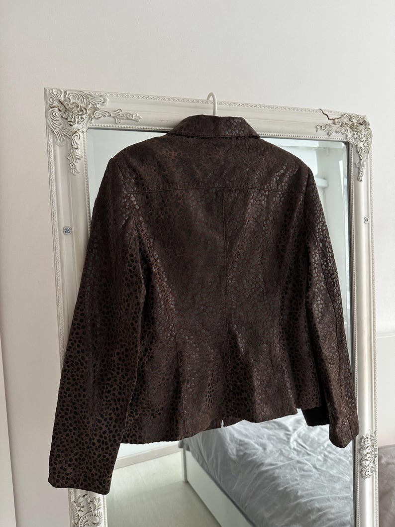 May include: A brown leather jacket with a textured, spotted pattern hangs on a hanger in front of a mirror. The jacket has a collared neckline and long sleeves. The mirror has a white ornate frame.