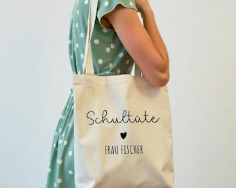 Könnte beinhalten: Eine cremefarbene Tragetasche mit den schwarzen Aufdrucken "Schultüte" und "FRAU FISCHER", sowie einem kleinen schwarzen Herz. Die Tasche hat cremefarbene Träger und wird von einer Person in einem grünen Polka-Dot-Kleid getragen.