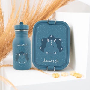 Könnte beinhalten: Ein teal-farbenes Wasserflaschen- und Lunchbox-Set. Beide Artikel zeigen ein Cartoon-Dinosaurier-Design und den Namen "Janosch" in weißer Schrift. Die Lunchbox hat eine rechteckige Form mit einem sicheren Verschluss. Die Flasche hat einen Schraubverschluss.