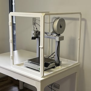 May include: A white 3D printer enclosed in a clear plastic enclosure. The printer is on a white table with a spool of filament on top of the enclosure.