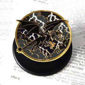 Könnte beinhalten: Eine kreisförmige Emaille-Anstecknadel mit der Silhouette eines schwarzen Drachen, der vor einem dunkelblauen Hintergrund mit weißen Blitzen und grauen Wolken fliegt. Das Design ist von einem goldfarbenen Metallrand umgeben und auf einem schwarzen Sockel.