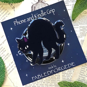 Pode incluir: Suporte para telefone e Kindle com um gato preto. O gato tem uma coleira roxa e está em um fundo azul escuro. O texto "Phone and Kindle Grip" está acima do gato. Fabricado por Fabledforgede.