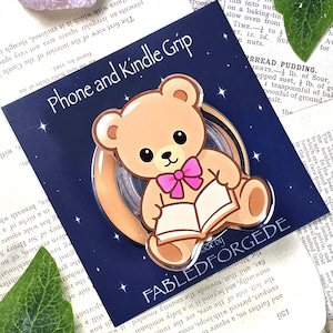 Peut inclure: Un support pour téléphone et Kindle avec un ours en peluche de dessin animé lisant un livre, avec un nœud papillon rose. Le support est sur fond bleu marine avec des étoiles, et le texte "FABLED FORGEDE".