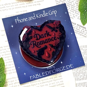 Peut inclure: Un support de téléphone en forme de cœur avec l'inscription "Dark Romance" en police gothique. Le support est noir et rouge, avec des roses, des pistolets et des larmes. Le support est sur un fond bleu.