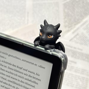 Könnte beinhalten: Eine kleine schwarze Drachenfigur mit orangefarbenen Augen, die auf der oberen Ecke eines E-Readers sitzt. Der Drache hat kleine Hörner und Flügel und ist über dem Rand des Bildschirms positioniert, auf dem deutscher Text in Schwarz zu sehen ist.