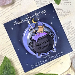 Pode incluir: Um suporte para telefone e Kindle com um caldeirão preto com poção roxa e o texto "Brewing Stories". O design inclui uma espada, um pergaminho, uma lua e estrelas sobre um fundo azul escuro.