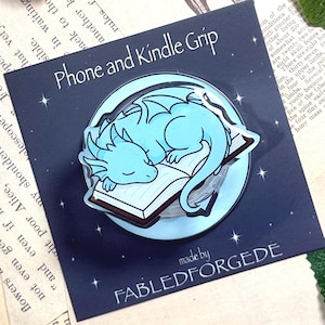 Könnte beinhalten: Ein Telefon- und Kindle-Griff mit einem hellblauen Drachen, der auf einem Buch schläft. Der Griff hat einen dunkelblauen Hintergrund mit dem Text "Phone and Kindle Grip" und "made by FABLEDFORGEDE". Das Design ist kreisförmig und hat ein glänzendes Finish.