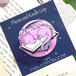 Könnte beinhalten: Ein Telefon- und Kindle-Griff mit einem schlafenden rosa Drachen auf einem Buch. Das Design befindet sich auf einem dunkelblauen Hintergrund. Der Text lautet "Phone and Kindle Grip" und "made by FABLEDFORGEDE".
