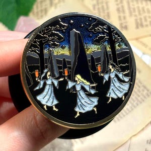 Op de afbeelding: Een zwarte en gouden ronde emaille pin met een nachtscène. Drie figuren in vloeiende gewaden dansen rond stenen. De achtergrond toont een sterrenhemel en bomen. De pin wordt door een hand vastgehouden.