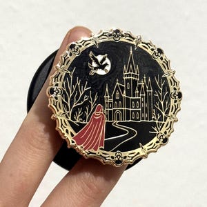 以下が含まれることがあります： 黒と金のゴシック様式の城のシーンが描かれた丸いエナメルピン。赤いマントを着た人物が城に向かって歩いています。ピンにはスカルのボーダーが付いています。