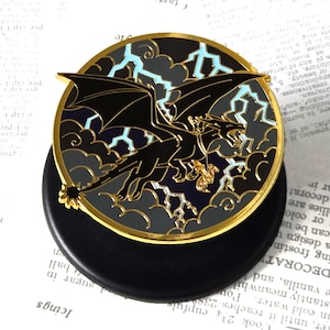 May include: A round, black enamel pin featuring a black dragon with gold accents, set against a backdrop of stormy clouds and lightning bolts. The pin has a gold border and is mounted on a black base.