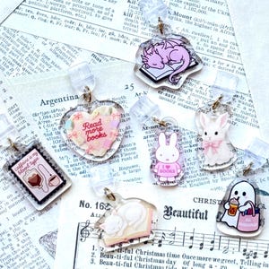 Puede incluir: Una colección de colgantes acrílicos transparentes con varios diseños, incluyendo un dragón rosa, un corazón con "Read more books", un conejito blanco y un fantasma con una bebida.