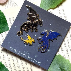 Könnte beinhalten: Drei Drachen-Emaille-Anstecknadeln in Schwarz, Gold und Blau mit goldenen Akzenten. Die Anstecknadeln befinden sich auf einer dunkelblauen Karte mit dem Text "FABLEDFORGEDE" und einem Sternenmuster. Fantasy-Thema.