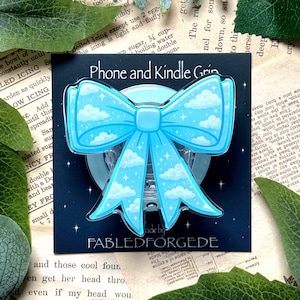 May include: A light blue phone and Kindle grip shaped like a bow, featuring a cloud design. The grip is set against a black backdrop, with the text "Phone and Kindle Grin" and "FABLEDFORGEDE".