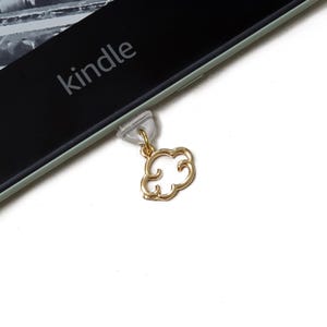 Op de afbeelding: Een goudkleurige wolkbedel bevestigd aan een doorzichtig plastic stuk, verbonden met een Kindle-apparaat. De Kindle heeft het woord "kindle" op het scherm gedrukt. De achtergrond is wit.