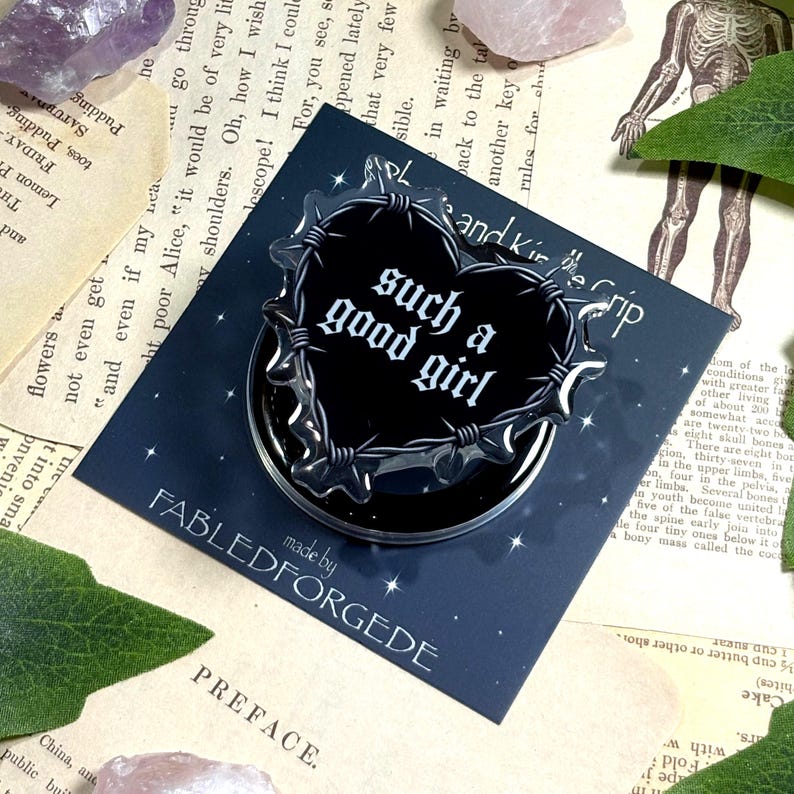 May include: A black heart-shaped pin with the text "such a good girl" in white gothic font, surrounded by a barbed wire design. The pin is on a dark blue background with the words "FABLEDFORGEDE" and "PREFACE" visible.
