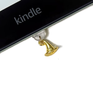 May include: A gold-coloured witch hat charm attached to a Kindle e-reader. The word "kindle" is visible on the black screen. The charm is suspended from a clear plastic piece.