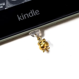 May include: A gold-coloured charm shaped like a small, stylised object, attached to a clear plastic piece. The charm is hanging from a Kindle device, with the word "kindle" visible on the screen.