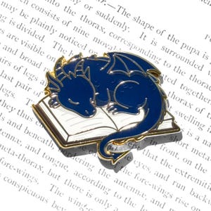 Könnte beinhalten: Dunkelblaue Drachenbrosche mit goldenen Rändern, die auf einem weißen Buch mit goldenen Kanten schläft. Der Drache hat detaillierte Flügel und einen eingerollten Schwanz. Ein Fantasy-Accessoire aus Metall.