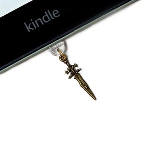 Puede incluir: Un dije de espada de color bronce, sujeto a un dispositivo Kindle. La palabra "kindle" está impresa en el dispositivo. El dije presenta un diseño detallado de empuñadura y hoja.