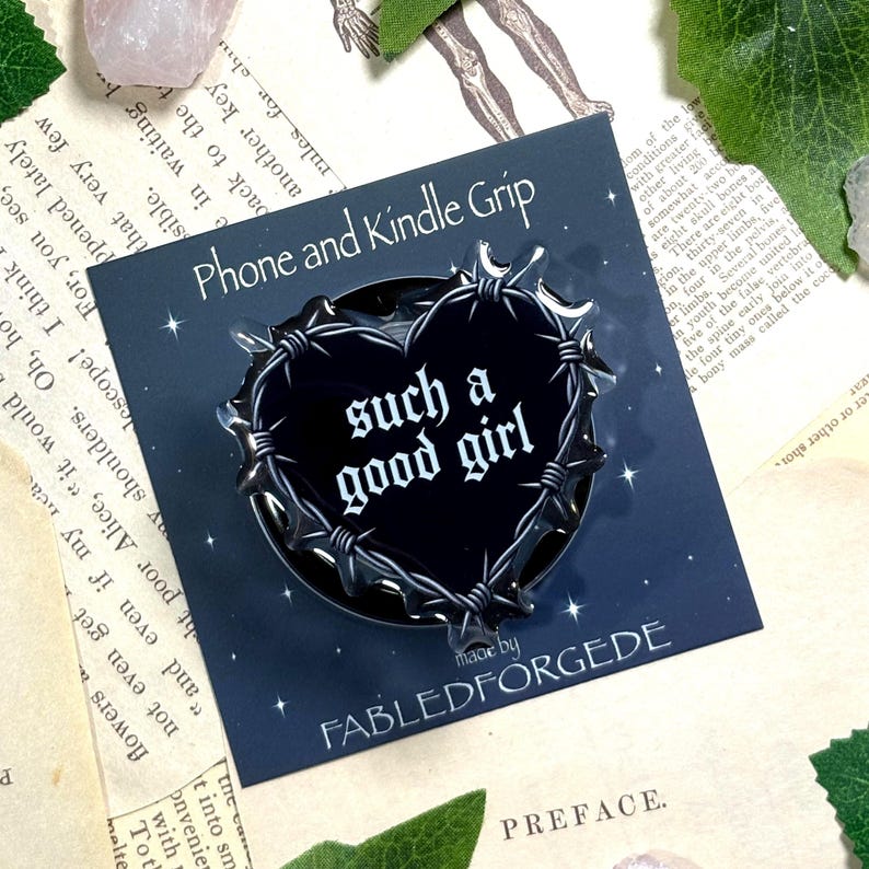 May include: A black heart-shaped phone and Kindle grip with barbed wire design. The text "such a good girl" is in white gothic font. The product is made by Fabled Forgede.
