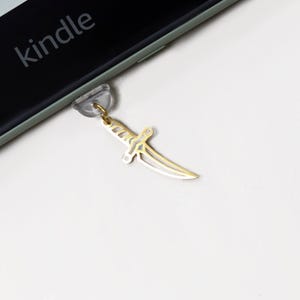 May include: A gold-coloured dagger charm attached to a clear suction cup. The charm is hanging next to a black Kindle device with the word "kindle" printed on the screen. The dagger has a detailed design.