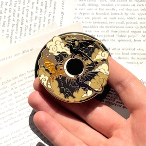 May include: A circular enamel pin featuring a central black circle surrounded by three dragons in flight. The design includes cream-coloured clouds, a gold glitter dragon, and two black dragons with gold outlines against a dark background.