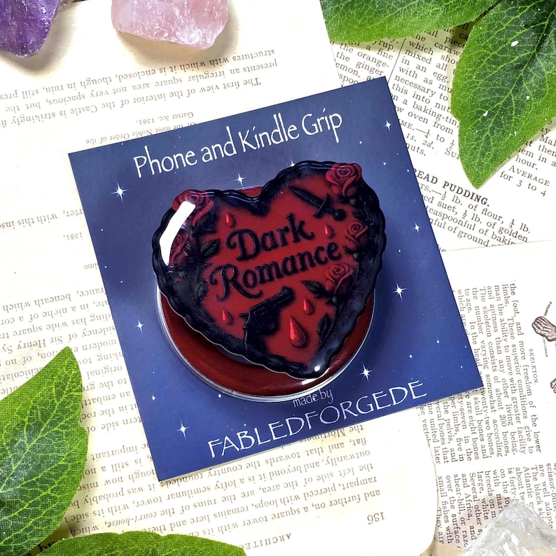 May include: A heart-shaped phone grip with the words "Dark Romance" in a gothic font. The grip is black and red, featuring roses, guns, and teardrop designs. The grip is on a blue background.