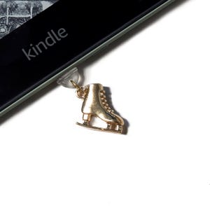 Könnte beinhalten: Ein goldfarbener Eislaufschuh-Anhänger. Der Anhänger ist an einem transparenten Kunststoffteil befestigt. Das Wort "kindle" ist auf einer schwarzen Oberfläche im Hintergrund zu sehen. Der Anhänger ist mit Schnürsenkeln und einer Klinge detailliert.