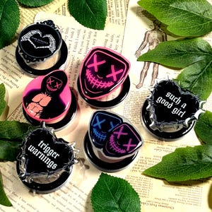 May include: A collection of black and clear phone grips with various designs. Some feature text like "Dark Romance" and "Trigger Warnings." Others display neon pink and blue mask graphics. The grips are arranged on a vintage paper background with green leaves.
