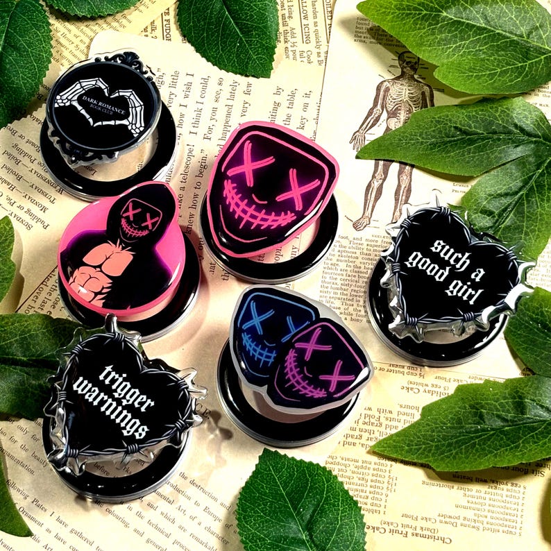 May include: A collection of black and clear phone grips with various designs. Some feature text like "Dark Romance" and "Trigger Warnings." Others display neon pink and blue mask graphics. The grips are arranged on a vintage paper background with green leaves.