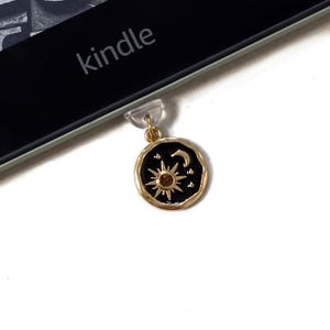 Puede incluir: Un dije dorado con fondo de esmalte negro, con un diseño de sol, luna y estrellas. El dije está unido a una pieza de plástico transparente, y la palabra "kindle" es visible en un dispositivo electrónico negro.