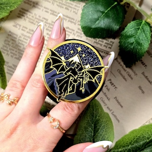 Könnte beinhalten: Runder Emaille-Pin in Schwarz und Gold, der eine geflügelte Silhouette vor einer sternenübersäten Berglandschaft zeigt. Der Pin wird von einer Hand mit goldenen Ringen und langen Nägeln gehalten.