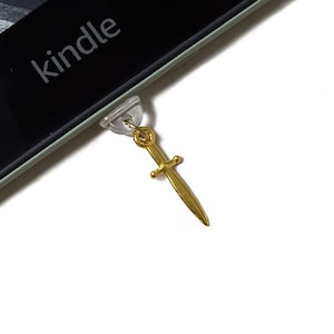 May include: A gold-coloured dagger charm attached to a clear plastic plug, designed to fit into an electronic device. The word "kindle" is visible on the device's screen. The charm is a decorative accessory.