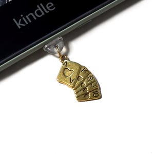 Puede incluir: Un dije dorado que representa un abanico de naipes, incluyendo el As de Corazones, Rey, Reina y 10 de Corazones. El dije está sujeto a una pieza de plástico transparente, probablemente para un teléfono o lector electrónico. La palabra "kindle" es visible en el dispositivo.